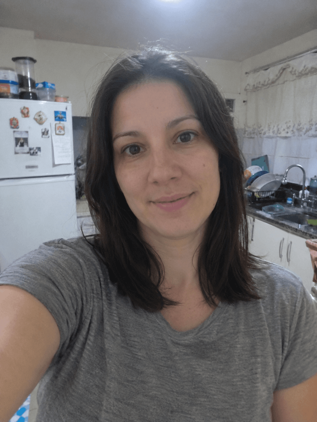 Selfie di una donna con t-shirt grigia in una cucina disordinata, luce da plafoniera