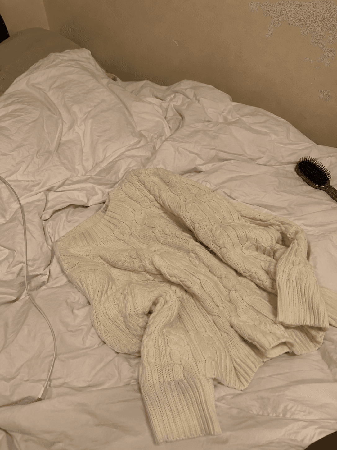 Maglione di lana color crema appoggiato sopra un letto disfatto, illuminazione artificiale