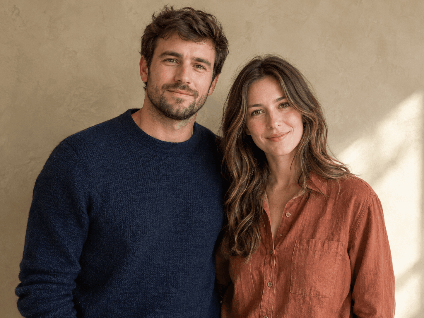 Portrait d’un homme en pull marine et d’une femme en chemise rouille côte à côte devant un mur beige