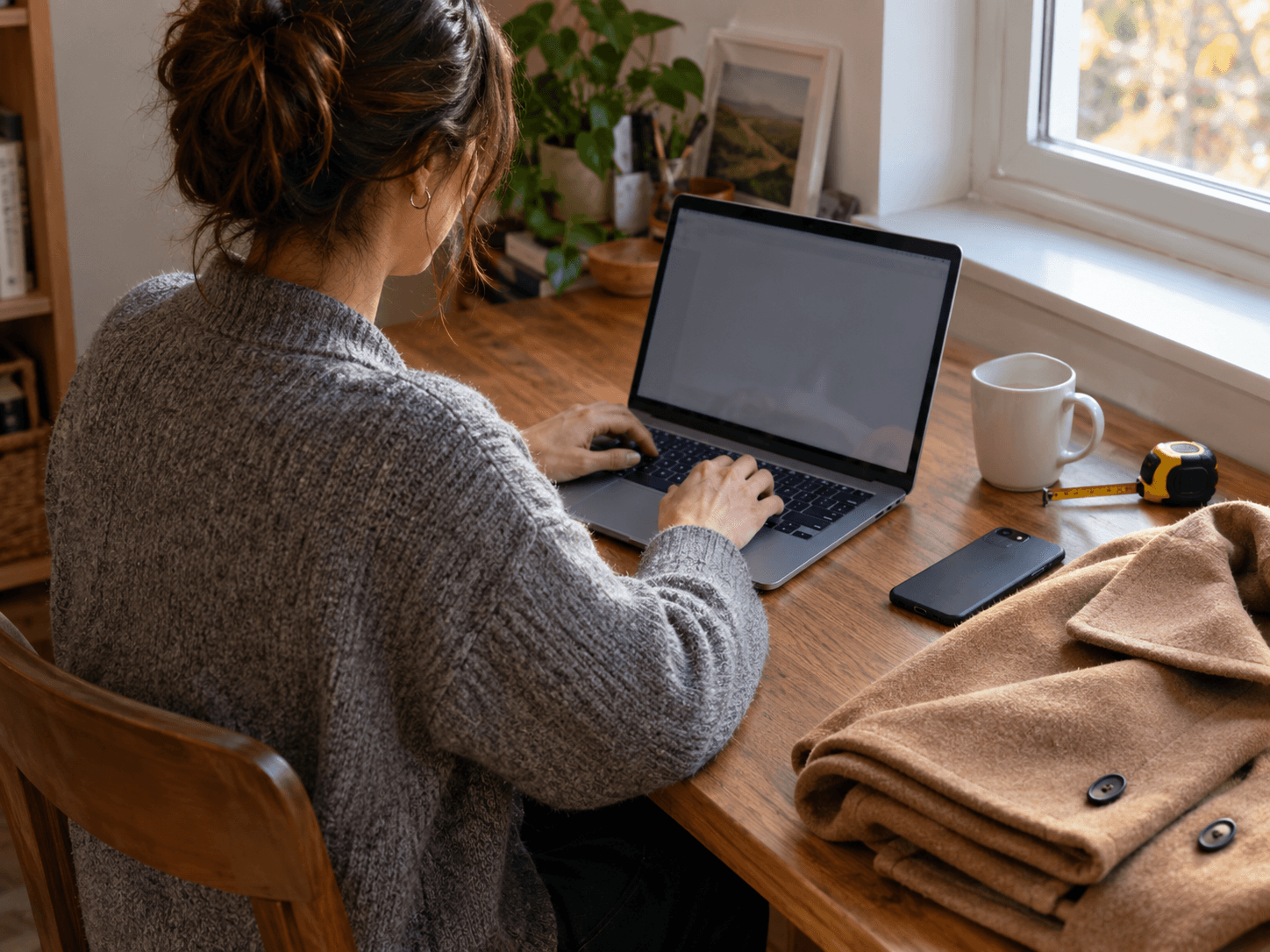 Frau im grauen Strickcardigan tippt am Laptop, daneben ein zusammengelegter Kamelmantel