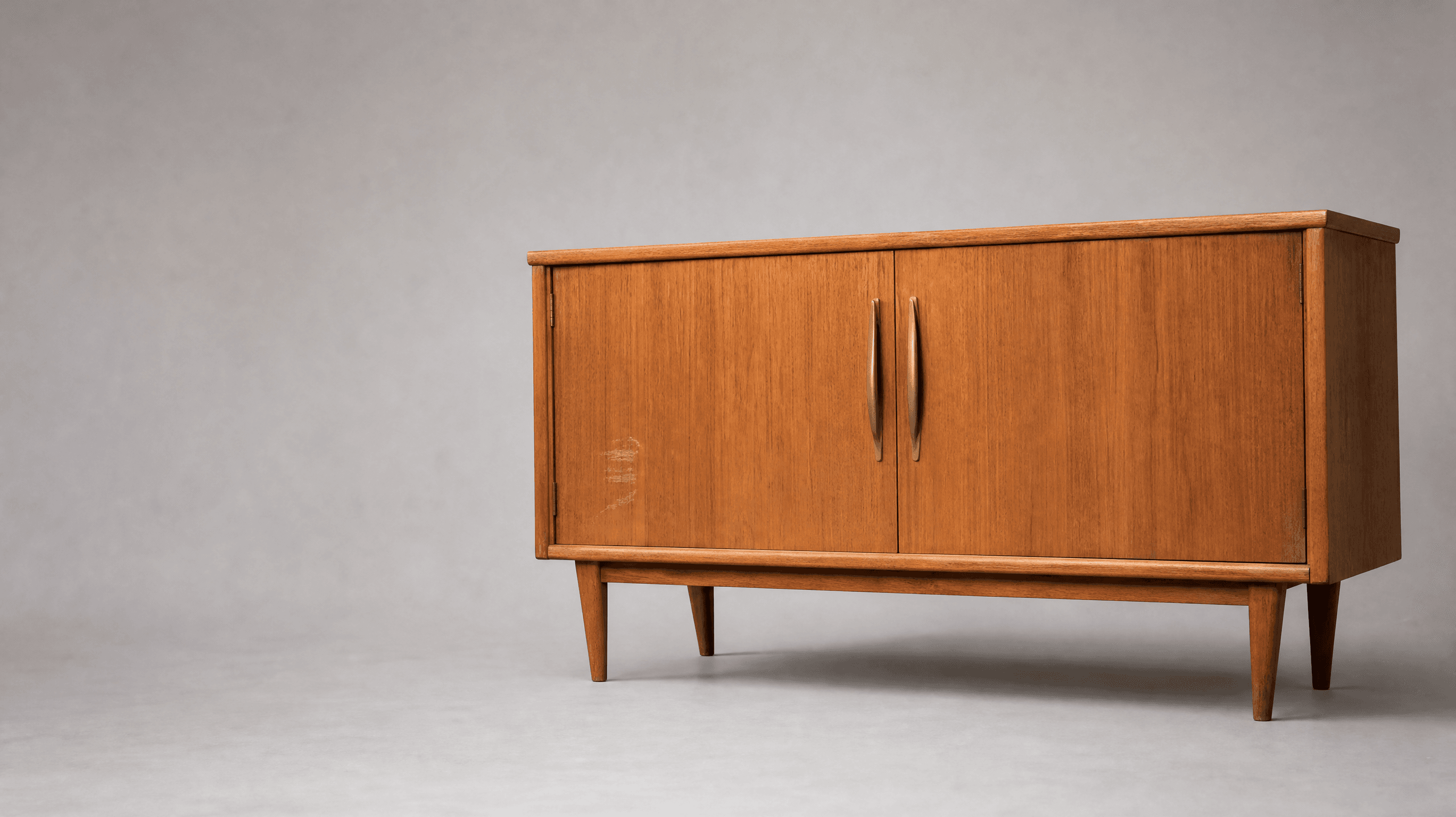 Gebrauchtes Teak-Sideboard mit zwei Messinggriffen vor hellgrauem Hintergrund, Kratzer an der linken Tür sichtbar