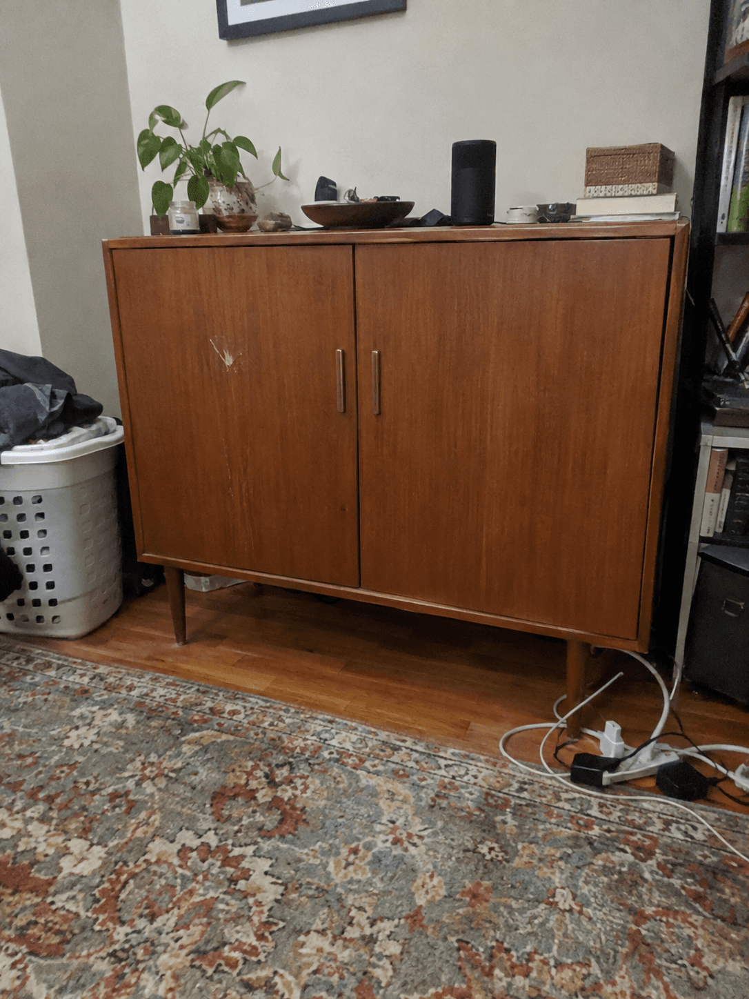 Handyfoto eines Teak-Sideboards vor einer vollgestellten Wohnzimmerwand mit Wäschekorb und Kabel am Boden