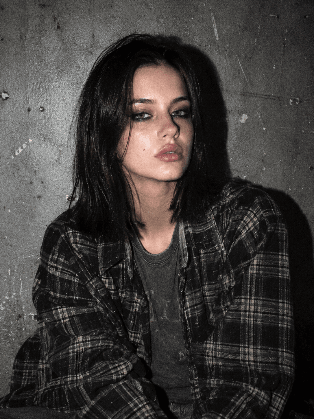 The same woman in a flannel shirt against a concrete wall, hard flash, shadow and grain