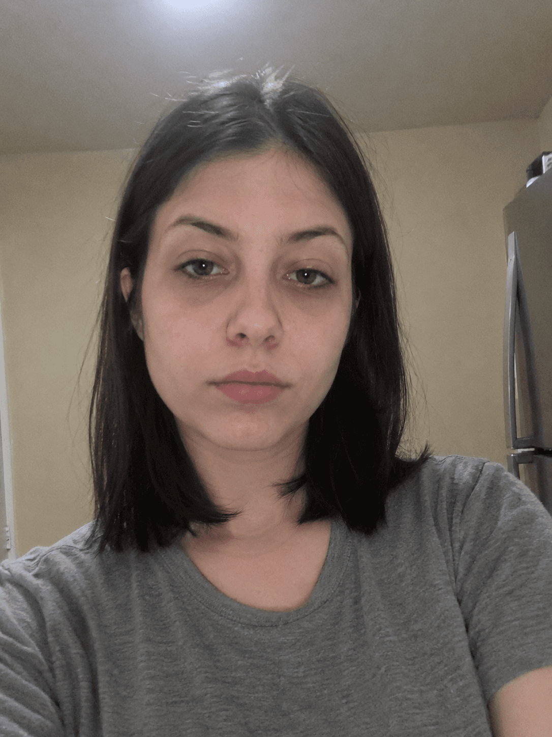 Plain selfie of a young woman under kitchen ceiling light, grey t-shirt, beige wall